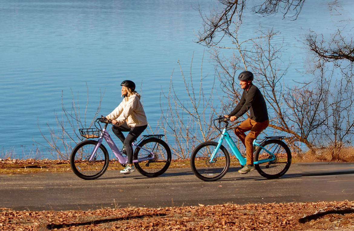 Two riders on a lakeside path on Velotric Breeze 1 e-bikes in Violet Haze and Ocean Mist, fitted with optional rear racks and a front basket, sold separately (Velotric photo) — Zeus eBikes Canada