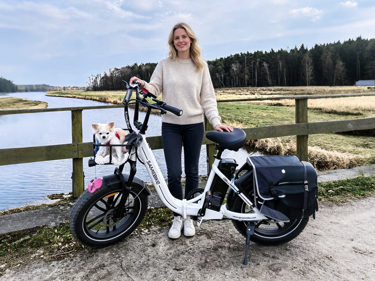 Rider standing beside a white Samebike C05 Pro by the water, a small dog in the front basket and a pannier bag on the rear rack; the bag is not included (Samebike's photo)
