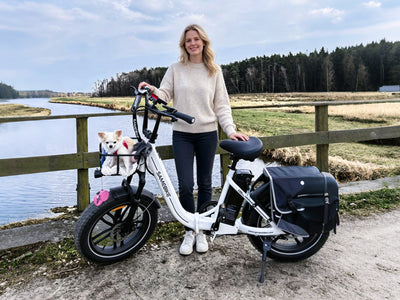 Rider standing beside a white Samebike C05 Pro by the water, a small dog in the front basket and a pannier bag on the rear rack; the bag is not included (Samebike's photo)