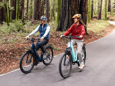 Two riders on Velotric Discover 3 e-bikes on a paved road through a forest (Velotric photo) — Zeus eBikes Canada