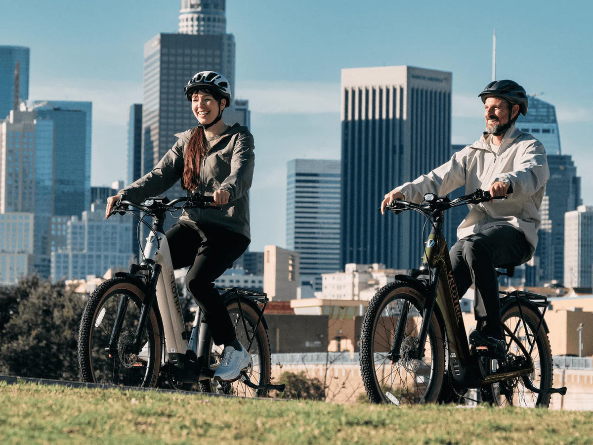Two riders on Velotric Discover M e-bikes with a city skyline behind them (Velotric photo) — Zeus eBikes Canada