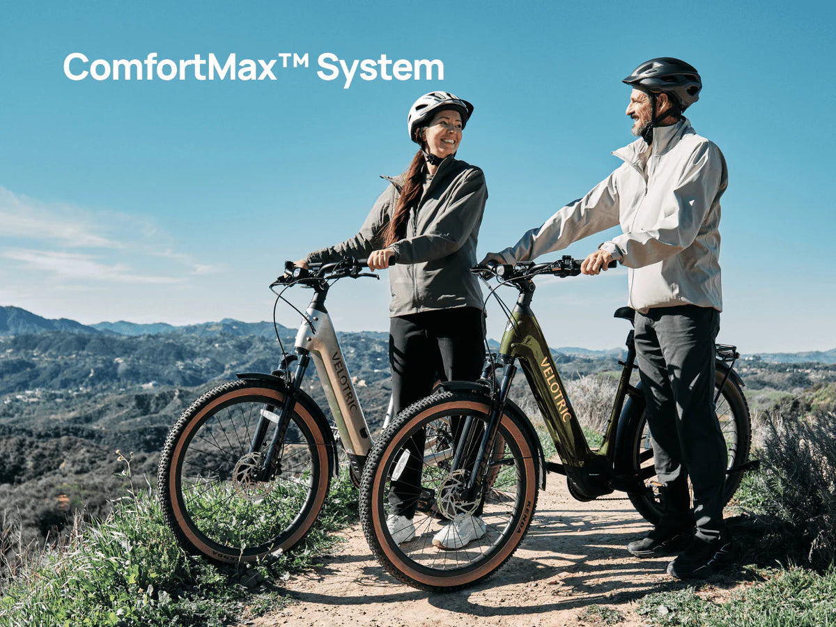 Two riders standing with Velotric Discover M e-bikes on a hilltop trail, with Velotric's ComfortMax caption — Zeus eBikes Canada